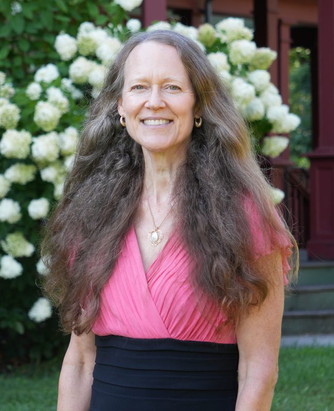 woman with long brown hair wearing dress smiling in front of flowering shrub